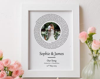 May include: A framed print with a white border features a wedding photo surrounded by song lyrics. The names "Sophie & James" are below the photo, with "Our Song" and the date beneath. A vase of pink roses sits nearby.