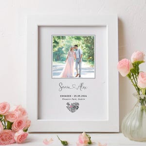 May include: A white picture frame with a photo of a couple in a park. The text "Sonia & Alex" is written in cursive above the photo. The text "ENGAGED - 25.05.2024" is written below the photo. The text "Phoenix Park, Dublin" is written below the date. A silver glitter heart is below the location.
