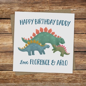 May include: A square greeting card with a brown backing. The card features a green Stegosaurus and two smaller dinosaurs in blue and green. Text reads "Happy Birthday Daddy" and "Love Florence & Arlo" in blue.