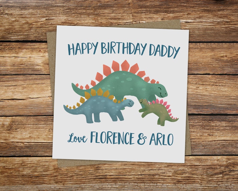 May include: A square greeting card with a brown backing. The card features a green Stegosaurus and two smaller dinosaurs in blue and green. Text reads "Happy Birthday Daddy" and "Love Florence & Arlo" in blue.