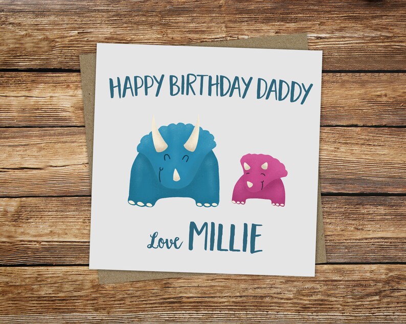 Puede incluir: Una tarjeta blanca con una ilustraci&oacute;n de dinosaurio azul y rosa. La tarjeta dice "Feliz cumplea&ntilde;os pap&aacute;" y "Con amor Millie".