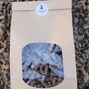 May include: A brown paper bag with a clear window showing brown wool fertiliser pellets. The bag has a white sticker with a red beet and the text "Wool Fertilizer Pellets".