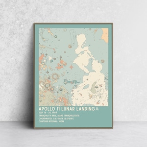 May include: A framed print featuring a map of the Apollo 11 Lunar Landing site. The artwork uses a muted color palette of blue, beige, and orange. Text at the bottom details the landing date, location, coordinates, and contour interval.