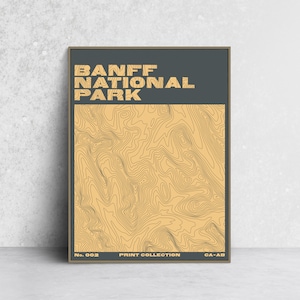 Peut inclure: Une affiche encadrée représentant une carte topographique du parc national de Banff. L'œuvre présente un fond bleu-vert foncé avec les mots "BANFF NATIONAL PARK" dans une police rétro. La carte est en tons de beige et de marron, avec le texte "No. 002 PRINT COLLECTION CA-AB" en bas.