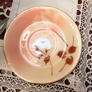 May include: A delicate pink saucer with a gold rim and a brown floral design. The saucer is made of porcelain and has a glossy finish.