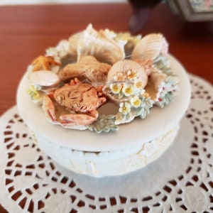 May include: A white ceramic trinket box with a seashell and crab design. The box is decorated with flowers and leaves.