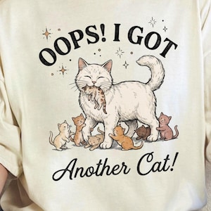 以下が含まれることがあります： クリーム色のTシャツ。猫のイラストが描かれています。大きな白い猫が子猫を口にくわえ、周りに小さな子猫がいます。「OOPS! I GOT Another Cat!」の文字が猫のイラストの上と下にアーチ状に配置されています。