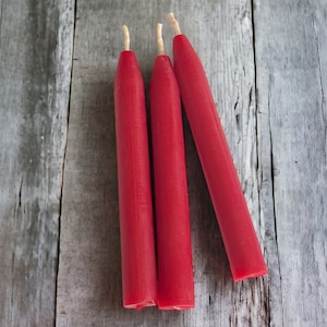 May include: Three red chime candles with  natural wicks on a weathered gray wooden surface.