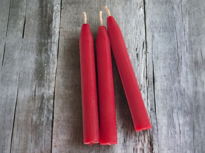 May include: Three red chime candles with  natural wicks on a weathered gray wooden surface.