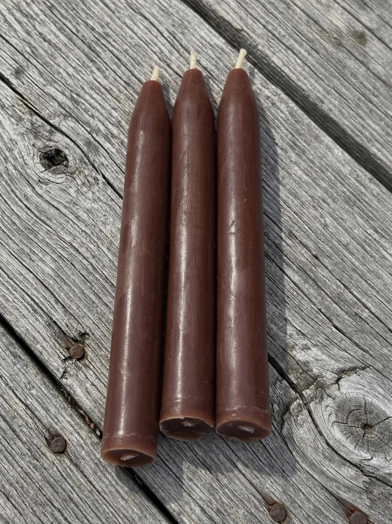 Pu&ograve; includere: Tre candele affusolate marroni con stoppini bianchi. Le candele sono cilindriche con una parte superiore leggermente affusolata. Le candele sono disposte verticalmente su una superficie di legno stagionato.