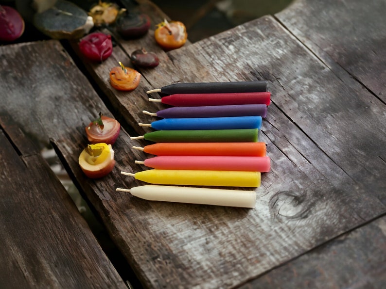 May include: A set of nine colorful candles on a wooden surface. The candles are arranged in a row from left to right, with the colors progressing from black to white. The colors of the candles are black, red, purple, blue, green, orange, pink, yellow, and white.