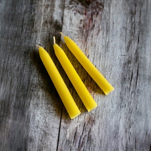 Pu&ograve; includere: Tre candele affusolate gialle con stoppini bianchi sono disposte diagonalmente su una superficie di legno grigio invecchiato. Le candele sono di un giallo vibrante e hanno una forma cilindrica liscia. Lo sfondo &egrave; in legno rustico e strutturato.