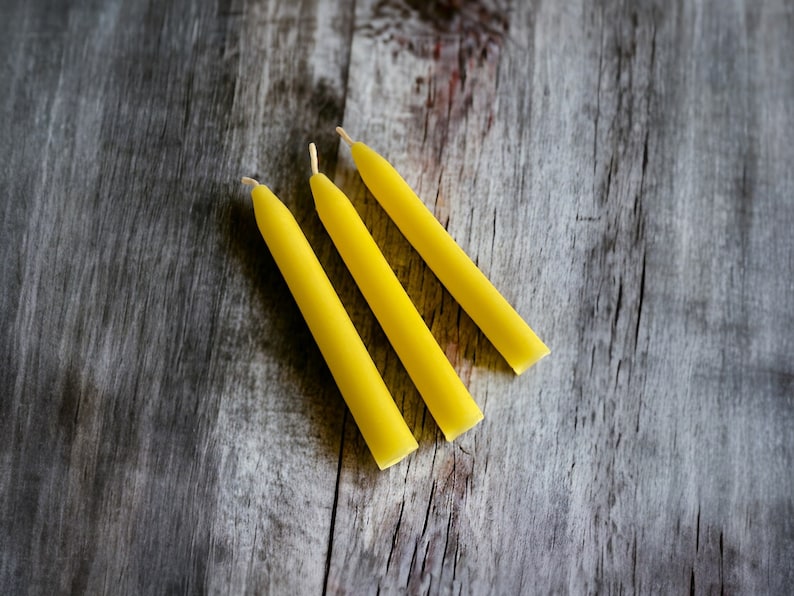 Pu&ograve; includere: Tre candele affusolate gialle con stoppini bianchi sono disposte diagonalmente su una superficie di legno grigio invecchiato. Le candele sono di un giallo vibrante e hanno una forma cilindrica liscia. Lo sfondo &egrave; in legno rustico e strutturato.
