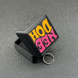 May include: A black, hinged box with the words "KEE DOH" in orange and pink lettering. The box is open, revealing a small interior and a silver-coloured keyring. The box appears to be 3D-printed.