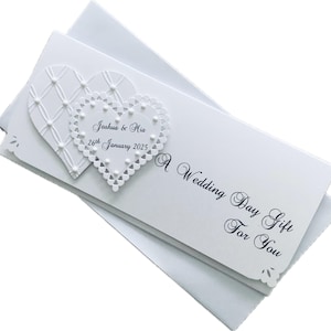 May include: A white wedding gift card with two heart-shaped embellishments. One heart has a lattice design, and the other has a scalloped edge with the names "Joshua & Mia" and the date "26th January 2025". The card reads "A Wedding Day Gift For You".