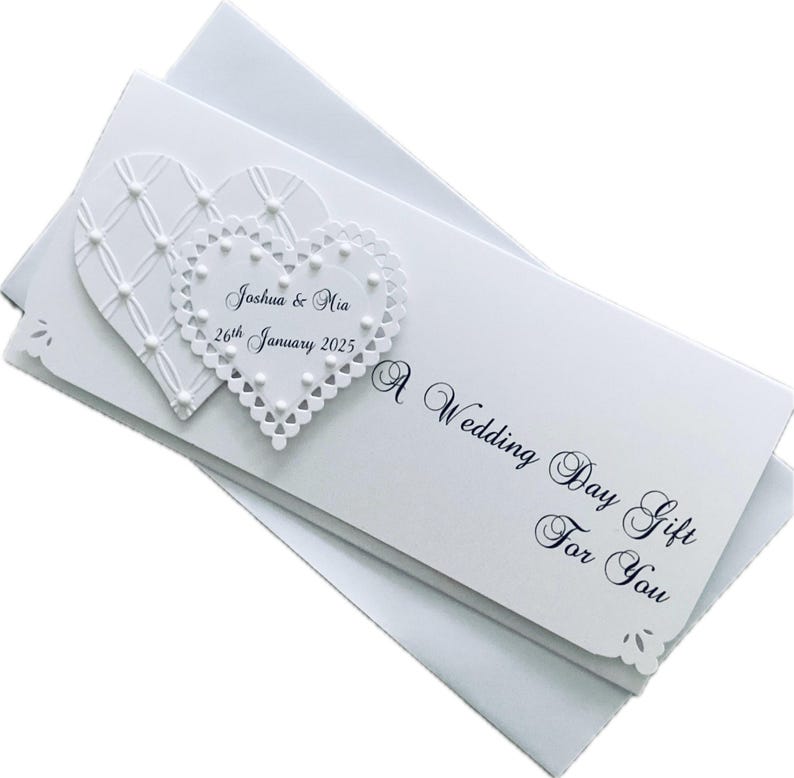 May include: A white wedding gift card with two heart-shaped embellishments. One heart has a lattice design, and the other has a scalloped edge with the names "Joshua & Mia" and the date "26th January 2025". The card reads "A Wedding Day Gift For You".