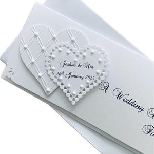 May include: White wedding invitation with two heart-shaped embellishments. One heart has a lattice design, and the other has a lace edge. The invitation includes the names "Joshua & Mia" and the date "26th January 2025".