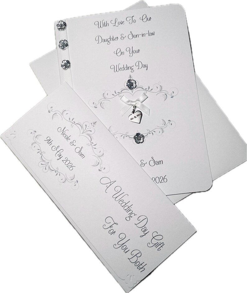 May include: A white wedding card set with silver script and embellishments. The main card reads "With Love To Our Daughter & Son-in-law On Your Wedding Day." A smaller card says "A Wedding Day Gift For You Both."