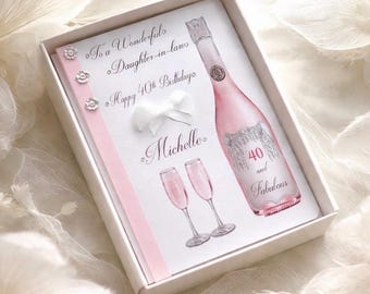 May include: A white gift box containing a greeting card with a pink champagne bottle illustration. The card reads "To a Wonderful Daughter-in-laws, Happy 40th Birthday Michelle, 40 and Fabulous". A white bow and pink ribbon accent the card.