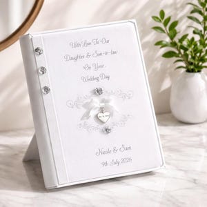 May include: A white wedding photo album with text that reads "With Love To Our Daughter & Son-in-law On Your Wedding Day". The album has decorative embellishments and a heart-shaped charm. The names "Nicole & Sam" and the date "9th July 2026" are also printed.