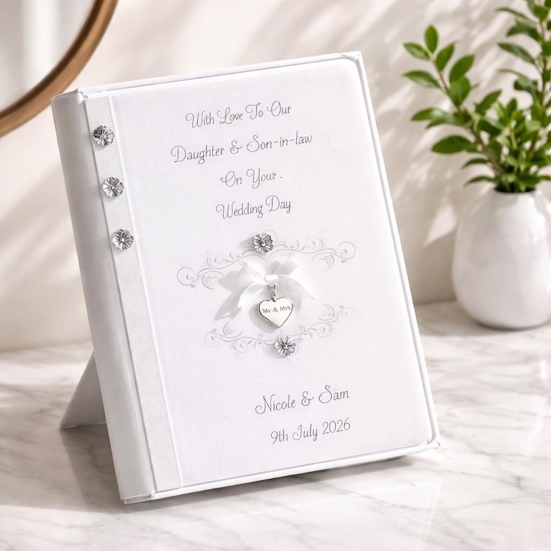 May include: A white wedding photo album with text that reads "With Love To Our Daughter & Son-in-law On Your Wedding Day". The album has decorative embellishments and a heart-shaped charm. The names "Nicole & Sam" and the date "9th July 2026" are also printed.