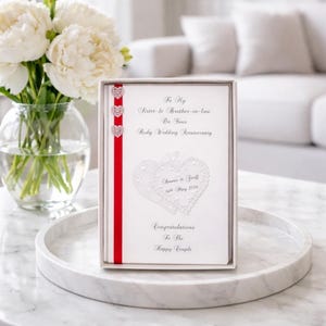 May include: A white wedding anniversary card in a white box, with a red ribbon and three heart-shaped embellishments. The card reads "To My Sister & Brother-in-law On Your Ruby Wedding Anniversary". A bouquet of white flowers is visible.