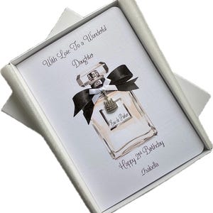 May include: A white greeting card in a white box, featuring a perfume bottle illustration with a black bow and a charm. The card reads "With Love To a Wonderful Daughter" and "Happy 21st Birthday Isabella."