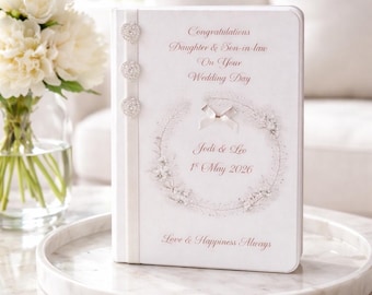 Könnte beinhalten: Ein weißes Hochzeitsgästebuch mit dem Text "Glückwunsch an Tochter & Schwiegersohn an Ihrem Hochzeitstag". Das Buch hat ein florales Kranzdesign und die Namen "Jodi & Leo" mit dem Datum "1. Mai 2026". Es ist mit drei Schmucksteinen verziert.