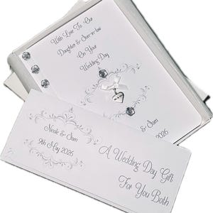 May include: A white wedding card set in a silver box. The card features decorative silver flourishes, a ribbon with a heart charm, and the text "With Love To Our Daughter & Son-in-law On Your Wedding Day". A separate card reads "A Wedding Day Gift For You Both".