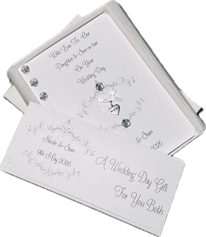 May include: A white wedding card set in a silver box. The card features decorative silver flourishes, a ribbon with a heart charm, and the text "With Love To Our Daughter & Son-in-law On Your Wedding Day". A separate card reads "A Wedding Day Gift For You Both".