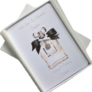 May include: A white greeting card with a perfume bottle illustration, black bow, and silver charm, inside a white box. Text reads "With Love To a Wonderful Daughter", "Happy 21st Birthday", and "Isabella".