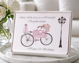 Puede incluir: Un juego de tarjetas de felicitación en una caja blanca. El diseño de la tarjeta presenta una bicicleta rosa que transporta flores y regalos, con el texto "Sent With Love to a Wonderful Daughter-in-law" y "Happy 50th Birthday."