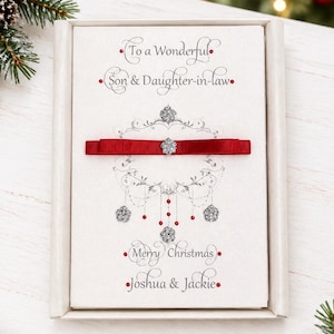 May include: A rectangular white greeting card in a box featuring the text To a Wonderful Son & Daughter-in-law and Merry Christmas Joshua & Jackie. The card has a red ribbon across the center with a silver flower accent and silver filigree.