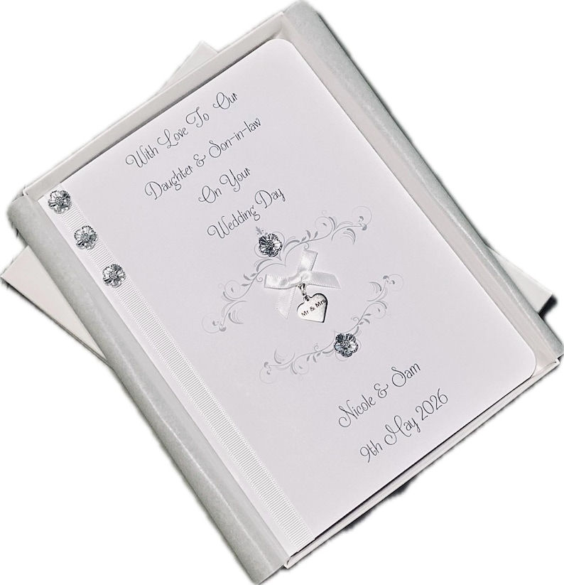 May include: A white wedding card with silver text and embellishments, presented in a white box. The card reads "With Love To Our Daughter & Son-in-law On Your Wedding Day" and includes the names "Nicole & Sam" and the date "9th May 2026".