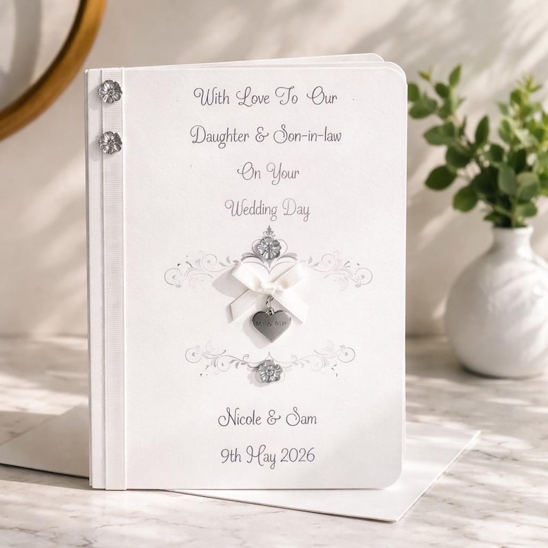 May include: A white wedding card with the text "With Love To Our Daughter & Son-in-law On Your Wedding Day". It features a silver heart charm and decorative embellishments. The names "Nicole & Sam" and the date "9th May 2026" are also printed on the card.