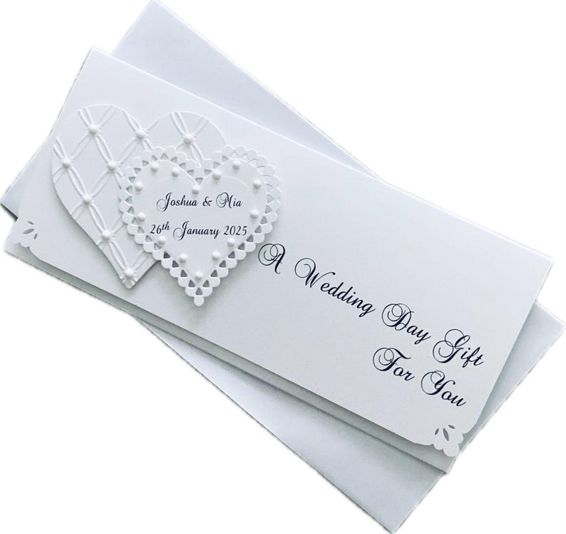 May include: White wedding day gift card with two heart-shaped embellishments. The card reads "Joshua & Mia 26th January 2025" and "A Wedding Day Gift For You". The card has a decorative edge and is placed on top of a white envelope.