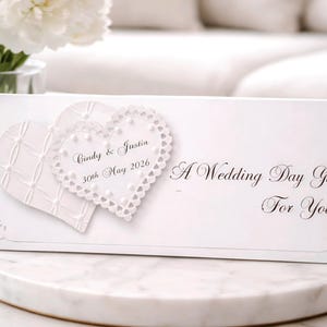 May include: A white wedding gift box with two heart-shaped embellishments. One heart has a textured pattern, and the other is a lace-edged heart. The box is inscribed with "Cindy & Justin 30th May 2026" and "A Wedding Day Gift For You."