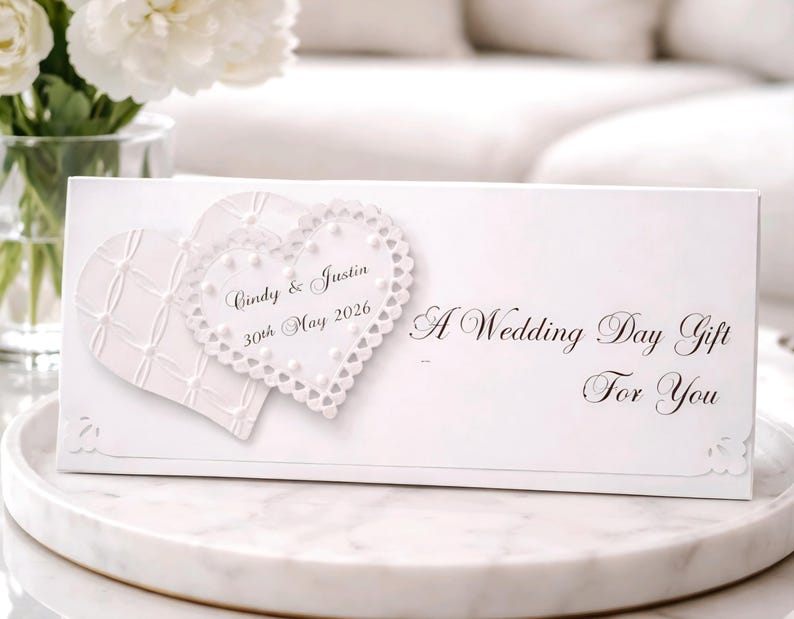 May include: A white wedding gift box with two heart-shaped embellishments. One heart has a textured pattern, and the other is a lace-edged heart. The box is inscribed with "Cindy & Justin 30th May 2026" and "A Wedding Day Gift For You."