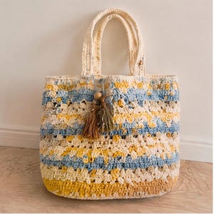 May include: A crochet tote bag featuring horizontal stripes in shades of cream, light blue, and yellow. The bag has two top handles and is decorated with two hanging tassels in brown and olive green, each accented with a round wooden bead.