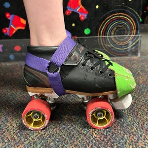 May include: Black roller skate with a green patch on the toe. The skate has a purple strap around the ankle and red wheels.