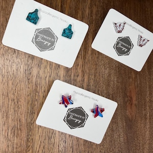 May include: Three pairs of earrings on white cards. One pair is teal with a swirl design, another is white with a cow face, and the last is red and blue star-shaped. Each card has "Hypoallergenic Hooks" and "HANDMADE BY Breezey" printed on it.