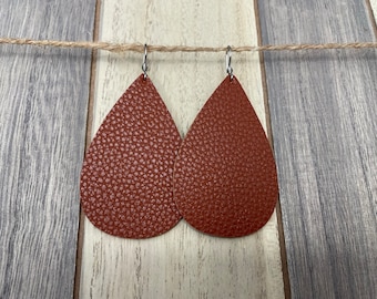 Peut inclure: Une paire de boucles d'oreilles en forme de goutte d'eau, de couleur brun rougeâtre. Les boucles d'oreilles ont un aspect texturé, semblable à du cuir, et sont suspendues à des crochets argentés. Elles sont présentées sur un fond de planches de bois.