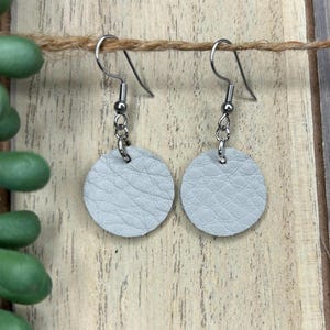 Puede incluir: Un par de pendientes de cuero gris claro con ganchos y detalles de cadena plateados. Los pendientes son redondos y tienen una superficie texturizada. El fondo incluye una superficie de madera y una planta verde.