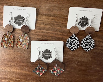 May include: Three pairs of earrings with hypoallergenic hooks. One pair features a floral design, another has a cow print, and the third has a mushroom pattern. Each earring set includes a wooden circle and a decorative acrylic shape.