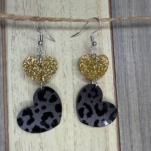 May include: A pair of dangle earrings featuring a gold glitter heart shape above a gray and black leopard print heart. The earrings hang from silver-colored hooks and are displayed against a wooden background.