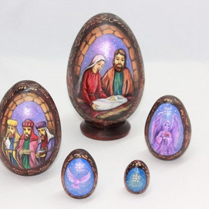 Peut inclure: Ensemble de cinq œufs de Noël en bois, chacun peint avec une scène différente de la Nativité. Le plus grand œuf représente la Sainte Famille, le suivant représente les trois mages, et les trois autres représentent des anges.