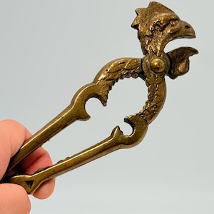 May include: A vintage brass nutcracker with a detailed eagle head design. The nutcracker features a lever-style mechanism and decorative curved handles. The brass has a warm, aged patina, suggesting it is an antique or vintage item.