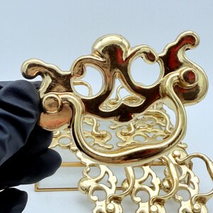 May include: Gold-coloured decorative drawer pulls with an ornate, swirling design. The pulls have a curved handle and decorative elements. The hardware is shown against a white background.