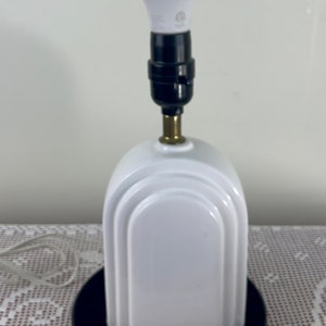 Può includere: Una lampada da tavolo in ceramica bianca con base nera e lampadina bianca. La lampada ha un design arrotondato di ispirazione Art Déco. Un righello di legno è posto vicino alla lampada.