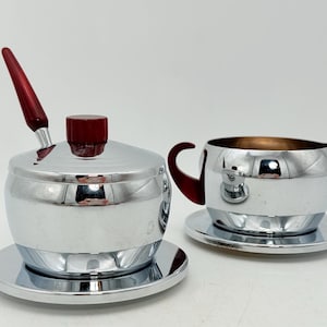 May include: A chrome sugar bowl with a red handle and lid, and a matching creamer with a red handle. Both pieces sit on small, round saucers. The set is designed for serving coffee or tea.